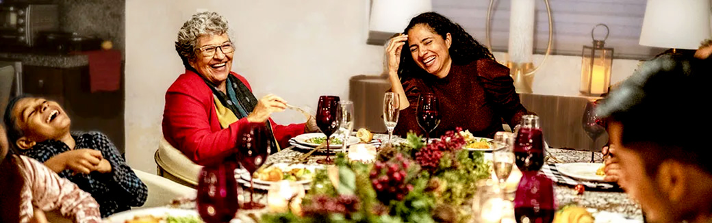 A multigenerational stepfamily gathers around a beautifully decorated dining table set with lots of holiday food, where they enjoy laughter along with their holiday dinner.