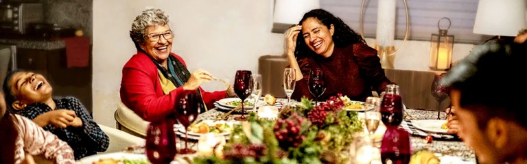 A multigenerational stepfamily gathers around a beautifully decorated dining table set with lots of holiday food, where they enjoy laughter along with their holiday dinner.