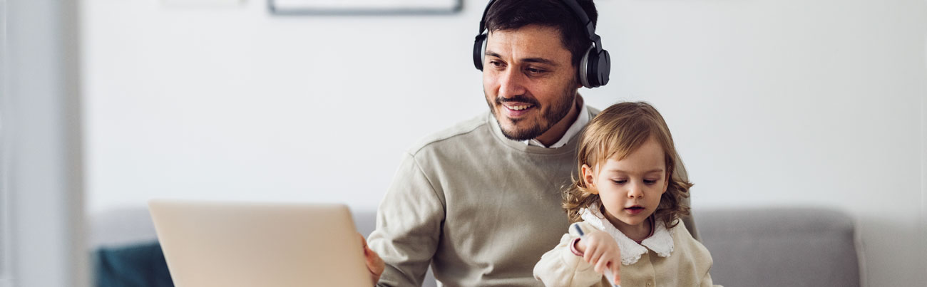 A father is balancing family time and his job as he works at his desk with a laptop at home, while his little daughter sits on his lap and pretends to be working alongside him, scribbling something in notebook.