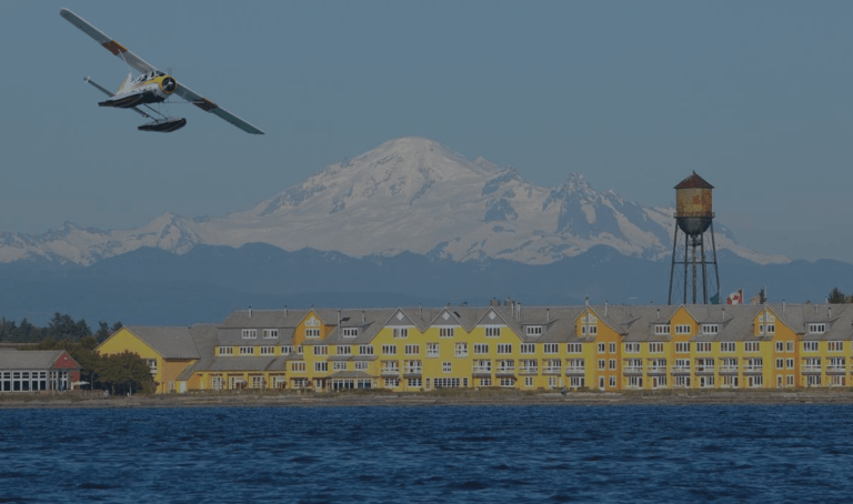 With the prominent snow-capped Mount Baker and city water tower in the background, a seaplane takes off with a portside view toward the row of yellow buildings lining the shore of the Semiahmoo Bay and Drayton Harbor of Bellingham (Blaine), Washington, a location of FamilyLife's Weekend to Remember® marriage retreat event.