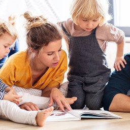 How To Create a Christ-Centered Family Culture A mother and father casually relaxing on the floor of their home with their two small children near them as they all intently look at a book together in an opportunity set up by the parents to help create a Christ-centered family culture