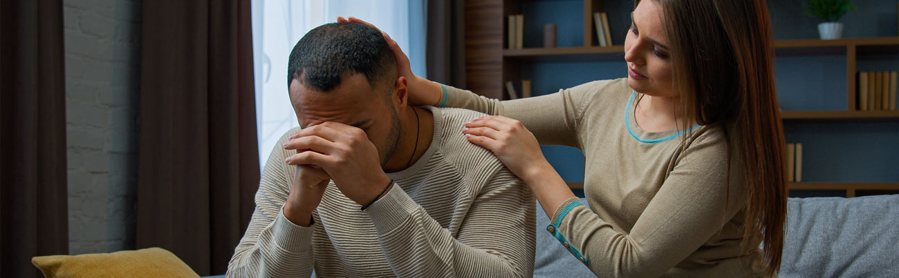 As they are seated in their home, a wife places her hands on her husband's shoulder while he is struggling to forgive her.