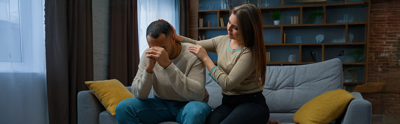 As they are seated in their home, a wife places her hands on her husband's shoulder while he is struggling to forgive her.
