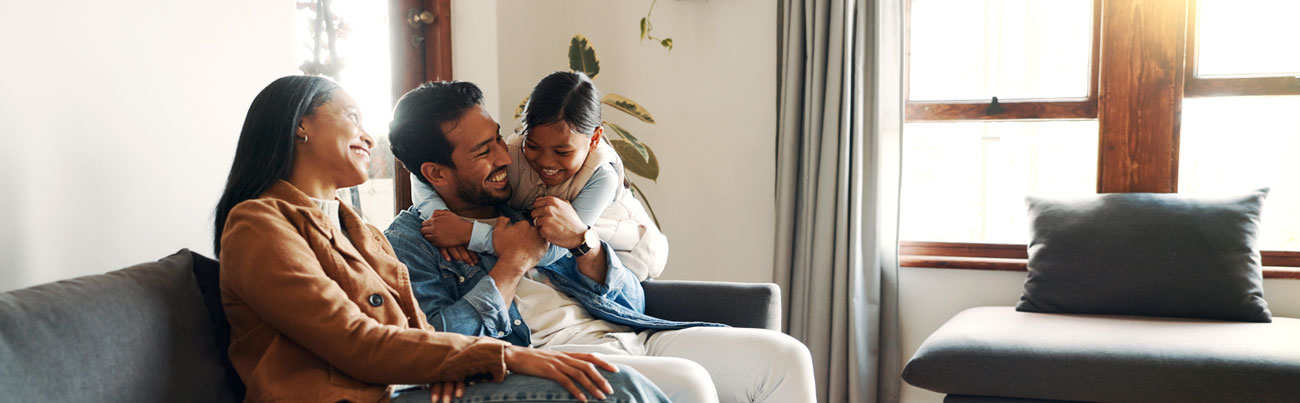 Smiling mother and father seated closely together on a sofa while their daughter joyously and with a big smile jumps up and wraps her arms around her father's neck after they finish a family talk about discipline