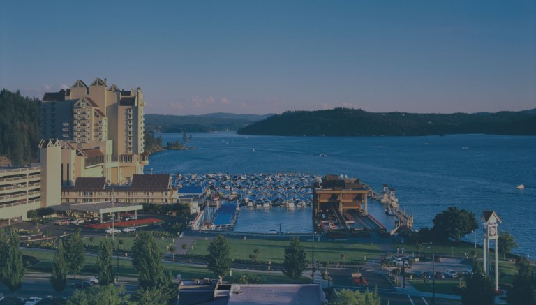 An expansive, bright daytime view showing off blue waters and hilltops in the background, and in the foreground The Coeur d'Alene Resort and marina, which is located directly on the shores of Lake Coeur d'Alene in downtown Coeur d'Alene, Idaho, a location of FamilyLife's Weekend to Remember® marriage retreat event.