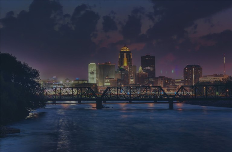 A view looking over the Des Moines River, and an old iron bridge in the foreground, with a clouded, stormy purple sky backlit by the sun as the backdrop for the downtown cityscape of Des Moines, Iowa, a location of FamilyLife's Weekend to Remember® marriage retreat event.