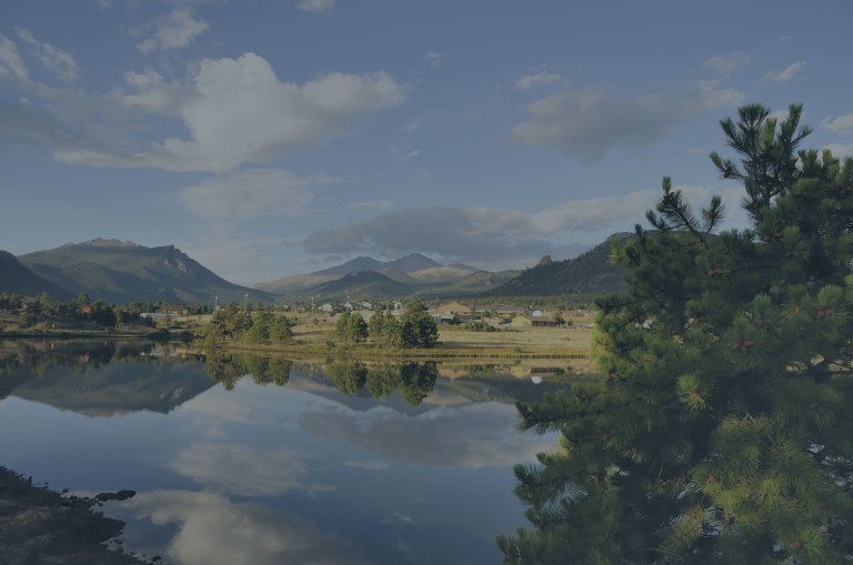 A dusky blue view overlooking the pristine, mirror-like surface of a lake, which is reflecting a portion of the Rocky Mountain National Park and white and gray clouds floating across the blue skies of Estes Park, Colorado, a location of FamilyLife's Weekend to Remember® marriage retreat event.