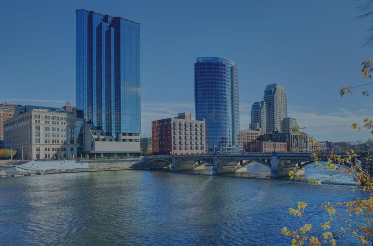 An across the water view with a backdrop of clear blue skies and an eye toward the cityscape of tall, modern, glass-covered buildings that line the banks of the Grand River, and a large bridge leads into the city of Grand Rapids, Michigan, a location of FamilyLife's Weekend to Remember® marriage retreat event.