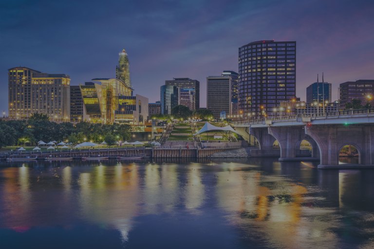 An early morning, lighted cityscape view reflecting onto the Connecticut River and the riverfront area along the downtown skyline, featuring Mortensen Riverfront Plaza, with grassy terraces, pedestrian walkways, and event stages that connect directly into the city of Hartford, Connecticut, a location of FamilyLife's Weekend to Remember® marriage retreat event.