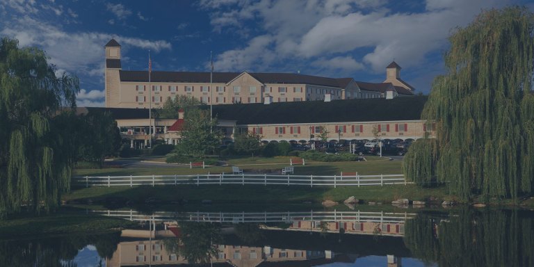 A picturesque landscape view of Hershey Lodge, flanked by two large weeping willow trees on the bank of the adjacent peaceful, still-water reflecting pond, located in Hershey, Pennsylvania, a location of FamilyLife's Weekend to Remember® marriage retreat event.
