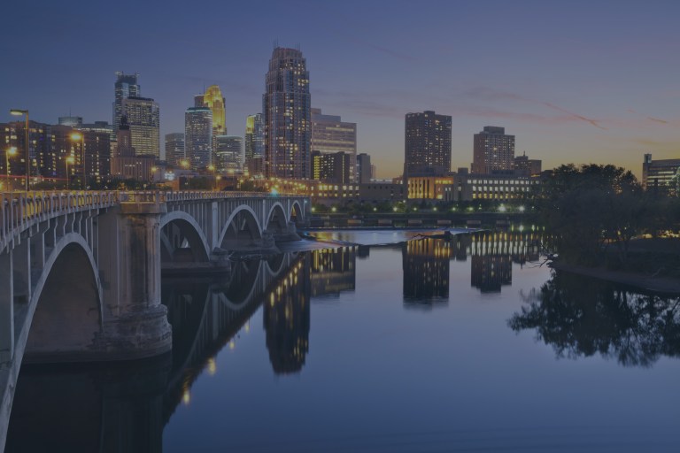 An early morning cityscape skyline view of the downtown lighted buildings lined up across the background, while the Stone Arch Bridge on the left crosses the Mississippi River in the foreground, and all of this scene is reflected in the river's waters, highlighting the beautiful city of Minneapolis, Minnesota, a location of FamilyLife's Weekend to Remember® marriage retreat event.