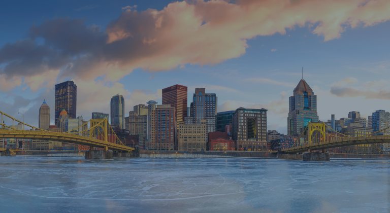 A daytime view between two expansion bridges overlooking the Allegheny River, with an eye toward the backdrop of blue skies partially covered with pastel tinted clouds, and the cityscape of buildings in downtown Pittsburgh, Pennsylvania, a location of FamilyLife's Weekend to Remember® marriage retreat event.