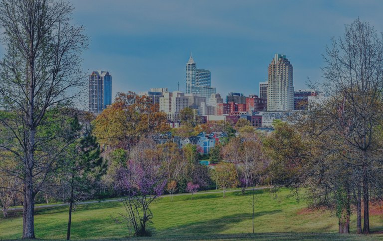 A city park with colorful trees and pedestrian pathways is situated in the forefront of this view, while an area of colorful residential homes line up behind the park, and the cityscape of tall buildings tower behind it all, making up the downtown area of Raleigh, North Carolina, a location of FamilyLife's Weekend to Remember® marriage retreat event.