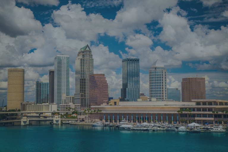 A view over the clear blue waters of Hillsborough Bay where it meets the edge of downtown streets, and where a cityscape of buildings rises on a background of blue skies with white clouds floating over Tampa, Florida, a location for FamilyLife's Weekend to Remember® marriage retreat.