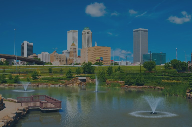 A pretty view across a landscaped pond with fountains, focusing your eye toward the downtown area and the horizon of city buildings that are set against a big blue sky in Tulsa, Oklahoma, a location for FamilyLife's Weekend to Remember® marriage retreat.