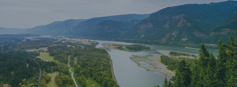 An aerial view over the flowing Colombia River with small islands in the middle and forest covered mountains on one side, all near the city of Vancouver, Washington, a location for FamilyLife's Weekend to Remember® marriage retreat.