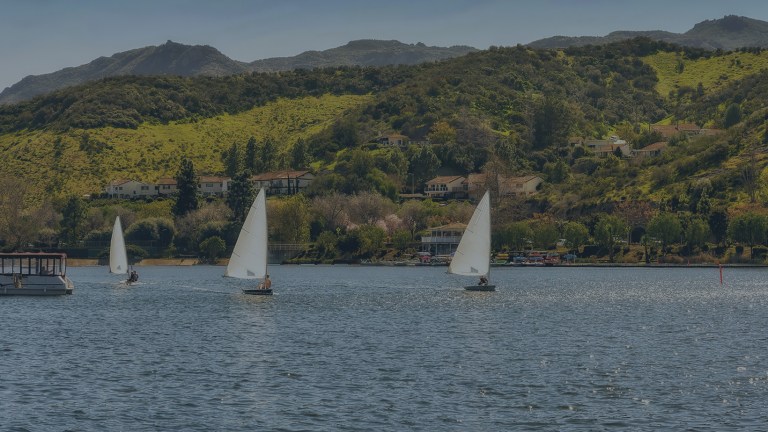 A view over Westlake Lake, a 125-acre man-made reservoir, where we see three small sailboats in the foreground and tree covered mountains as a backdrop to the city of Westlake Village, California, a location for FamilyLife's Weekend to Remember® marriage retreat.