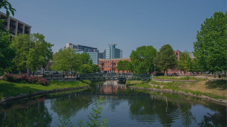 A view of some of the downtown city skyline in the background looking up from a view overlooking a beautifully landscaped, peaceful duckpond and footbridge at the center of Kalamazoo, Michigan, a location of FamilyLife's Weekend to Remember® marriage retreat event.