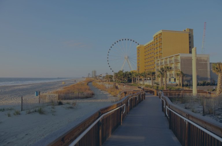 A white sand coastline ocean view toward the south, overlooking seagrass covered sand dunes, with a view down the wooden boardwalk meandering in front of the hotels, and the large Sky Wheel ferris wheel in the distance on Myrtle Beach, South Carolina, a location of FamilyLife's Weekend to Remember® marriage retreat event.