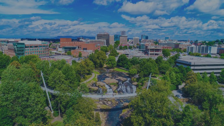 An aerial view of the downtown cityscape against blue clouded skies in the background, with the pedestrian Liberty Bridge in the foreground, spanning directly over the Reedy River, which flows over the stunning Reedy River Falls right beneath the bridge, all with the backdrop of the trees of Falls Park in Greenville, South Carolina, a location of FamilyLife's Weekend to Remember® marriage retreat event.