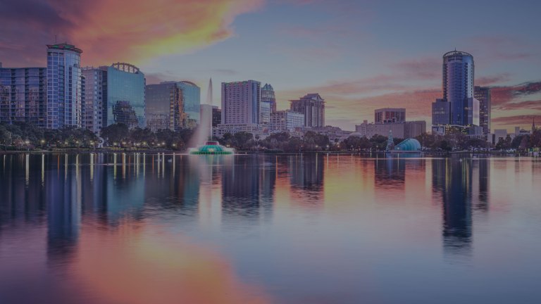Dusky blue skies with yellow and orange sun-tinged clouds reflect onto the centrally located Lake Eola and its iconic lighted fountain, with a backdrop of the lakeside city skyline of Orlando, Florida, a location of FamilyLife's Weekend to Remember® marriage retreat event.