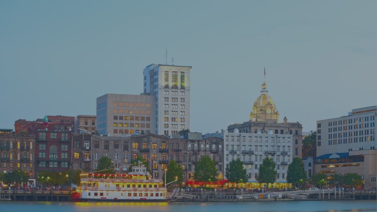 A cityscape skyline view from the Savannah River, which flows past an historic downtown and famous cobblestone-paved River Street, and shows off the marina along the bank, where a large red, yellow and white party riverboat is docked in front of downtown Savannah, Georgia, a location of FamilyLife's Weekend to Remember® marriage retreat event.