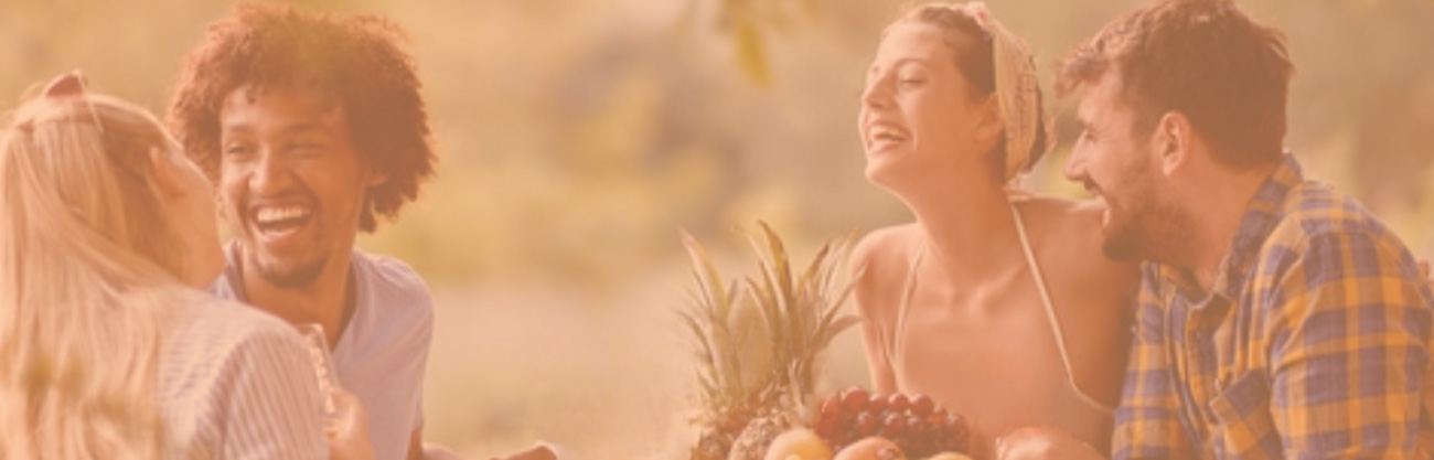 Two happy, laughing couples seated outdoors at a table under a tree with many types of fruit to enjoy.