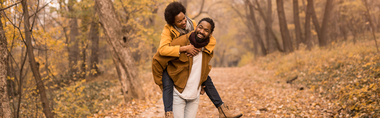 A happily married couple that keeps a list of fun fall date ideas, uses one from the list and playfully romps through the golden fall leaves along a wooded trail, as the husband carries his wife playfully in a piggyback style on his back.
